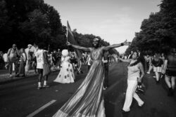 vogue 2 Eine Person mit langem Gewand trägt eine Regenbogenfarbe die Arme weit ausgetreckt. CSD 2023. Eine Frau mit einer Flasche in der Hand streckt die andere Hand zum Berühren aus
